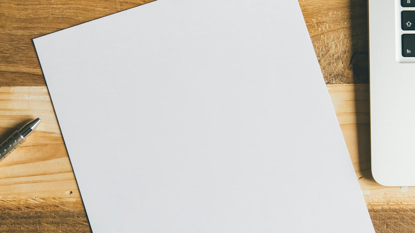Folio en blanco sobre una mesa de madera, junto a un bolígrafo y un portátil